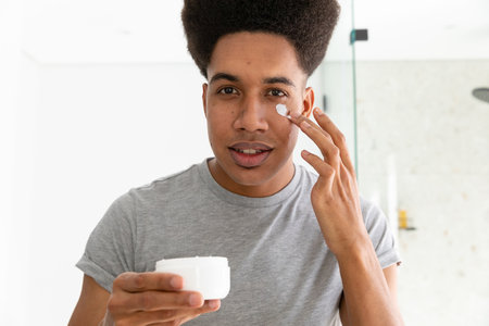 Biracial man looking in mirror and applying face cream in sunny bathroom, unaltered. Self care, skin care, wellbeing and lifestyle, unaltered.の写真素材