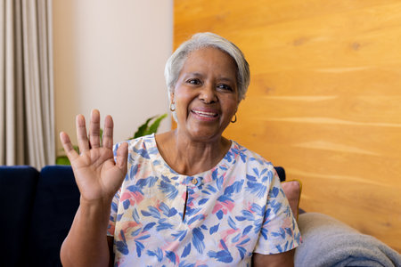 Happy senior biracial woman making video call smiling and waving sitting on sofa at home. Senior lifestyle, communication, technology and domestic life, unaltered.の写真素材