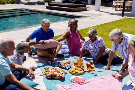 Happy senior diverse people sitting on blanket and having picnic in garden. Senior lifestyle, friendship and relaxation, retirement, unaltered.の写真素材