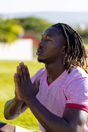 African american man sitting on grass, doing yoga and meditating in garden. Sport, healthy and active lifestyle, summer, sunshine, unaltered.の写真素材