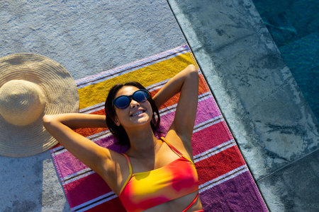 Happy biracial woman lying on towel and sunbathing next to swimming pool in garden. Lifestyle, free...