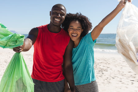 Portrait of happy african american couple cleaning sunny beach and collecting litter. Summer,...