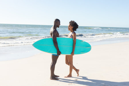 Happy african american couple holding surfboard smiling at each other standing on sunny beach. Summer, togetherness, romance, healthy lifestyle, sport, hobbies, surfing and vacation, unaltered.の写真素材