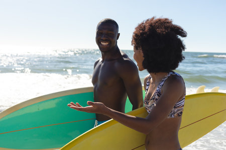 Happy, fit african american couple carrying surfboards talking, walking on sunny beach by the sea. Summer, healthy lifestyle, sport, hobbies, surfing and vacation, unaltered.の写真素材