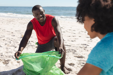 Happy african american couple cleaning sunny beach and collecting litter. Summer, lifestyle, free time, ecology, recycling and vacation, unaltered.の写真素材