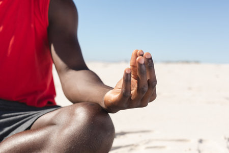 Midsection of fit african american man practicing yoga meditation sitting on sunny beach. Lotus position, summer, healthy lifestyle, relaxation, wellbeing and vacation, unaltered.の写真素材