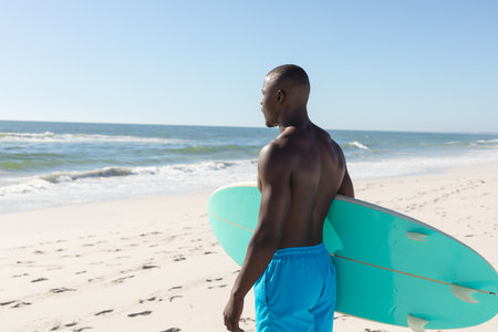 Fit african american man carrying surfboard on sunny beach, looking out to sea. Summer, healthy lifestyle, sport, hobbies, surfing and vacation, unaltered.の写真素材