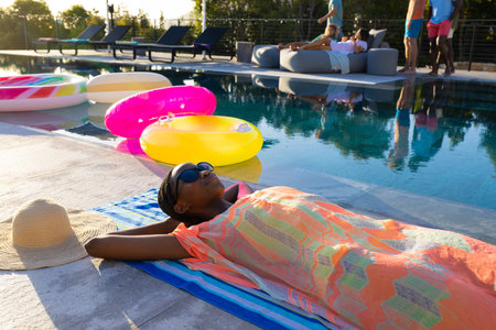 Happy african american woman lying on towel and sunbathing next to swimming pool in garden....