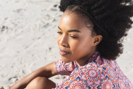 Happy african american woman smiling and looking away sitting on sunny beach. Summer, wellbeing,...