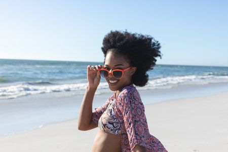 Portrait of happy african american woman in sunglasses smiling on sunny beach by sea. Summer, wellbeing, freedom, relaxation and vacation, unaltered.の写真素材
