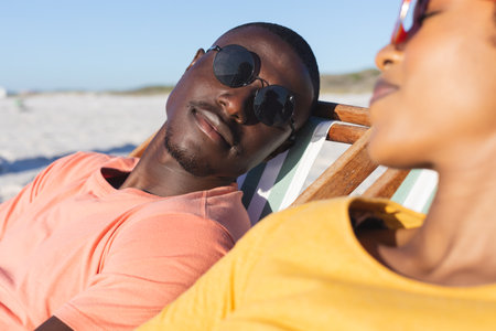 Happy african american couple in sunglasses sitting in deckchairs relaxing on sunny beach. Summer, romance, relaxation, togetherness and vacation, unaltered.の写真素材