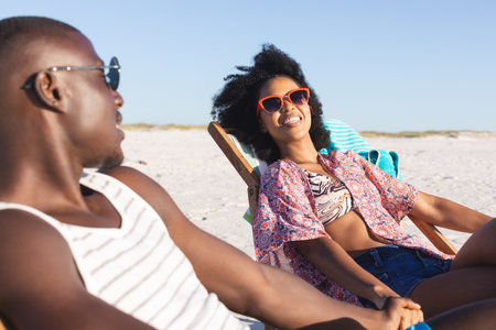 Happy african american couple sitting in deckchairs holding hands on sunny beach. Summer, romance, relaxation, togetherness and vacation, unaltered.の写真素材