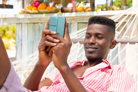 Happy biracial man lying in hammock using smartphone at sunny beach bar. Relaxation, communication, lifestyle, free time, summer and vacation, unaltered.の写真素材