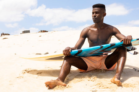 Focused biracial man holding surfboard sitting on sunny beach looking away. Active lifestyle,...