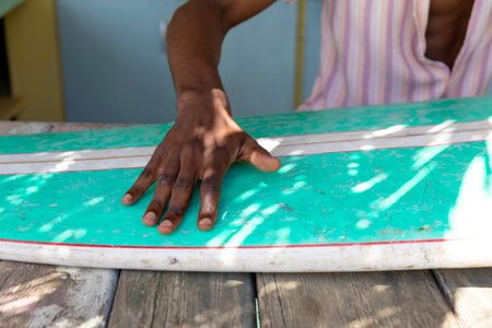 Midsection of african american man preparing surfboard behind counter of surf hire beach shack. Local business, business owner, hobbies, sport, surfing, summer and vacation.の写真素材