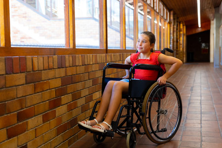 Happy caucasian schoolgirl sitting in wheelchair at elementary school corridor. Disability,...
