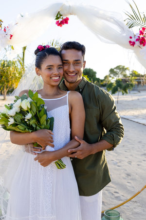 Smiling caucasian newlywed young couple with bouquet standing at beach at wedding ceremony. Copy space, unaltered, love, together, destination wedding, event, tradition, celebration, happy.の写真素材