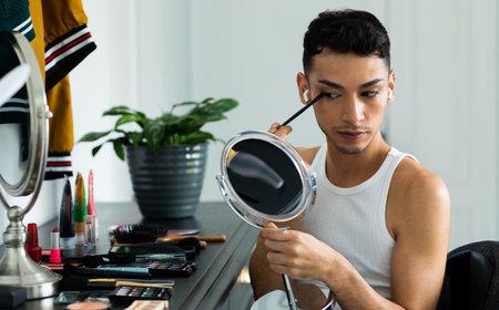 Biracial transgender man looking in mirror and putting on make-up, applying eyeshadow. Gender, self care, beauty, lifestyle and domestic life, unaltered.の写真素材