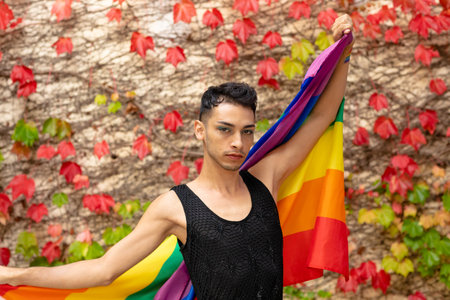Portrait of biracial transgender man holding rainbow flag, with rocks and leaves in background....