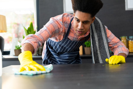 Focused biracial man wearing apron and rubber gloves cleaning countertop in sunny kitchen. Cleaning,...