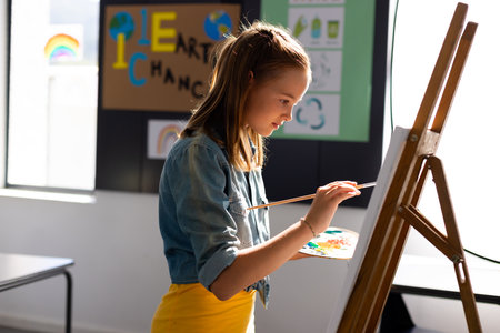 Happy caucasian schoolgirl painting using brush and easel in school art class. School, education,...