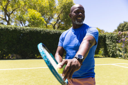 Happy senior african american man playing tennis preparing to serve on sunny grass court. Senior...