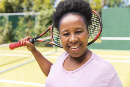 Portrait of senior african american woman with tennis racket smiling on sunny grass court. Senior lifestyle, retirement, sport, summer, fitness, hobbies and leisure activities.の写真素材