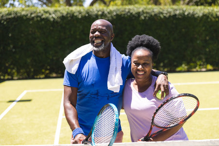 Happy senior african american couple with tennis rackets embracing on sunny grass court. Senior...