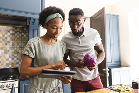 Happy african american couple preparing meal together using tablet in kitchen. Cooking, healthy lifestyle, recipe, communication, togetherness, food and domestic life, unaltered.の写真素材