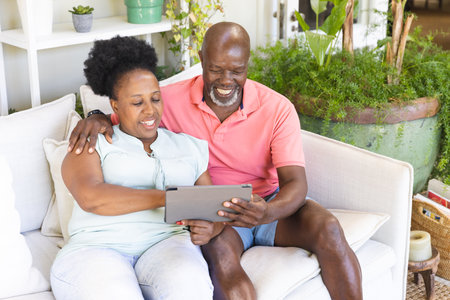 Happy senior african american couple on couch using tablet and embracing in sunny living room....