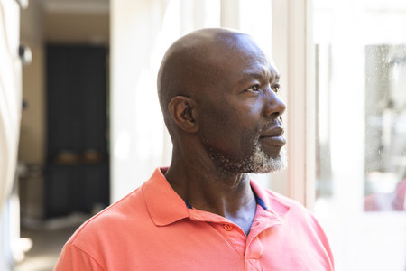Thoughtful, happy senior african american man standing looking out of window at home. Senior...