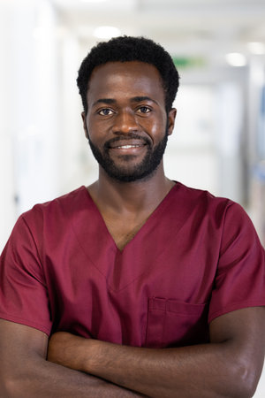 Unaltered portrait of african american male doctor in scrubs smiling, standing in hospital corridor....
