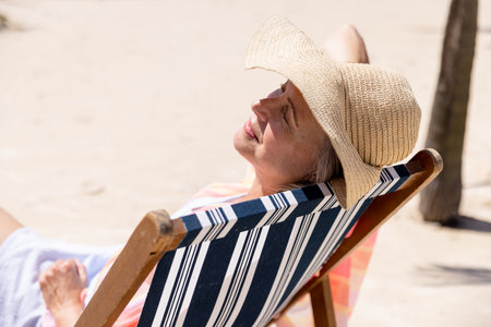 Caucasian senior woman wearing hat and with eyes closed sleeping on deck chair at beach. Copy space, unaltered, retirement, vacation, lifestyle, enjoyment, relaxation and summer concept.のeditorial素材