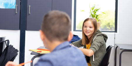 Happy caucasian schoolgirl talking with schoolboy in classroom, with copy space. Education, school...