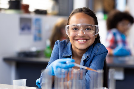 Portrait of happy biracial schoolgirl wearing safety glasses in chemistry class. Education, inclusivity, elementary school and learning concept.の写真素材