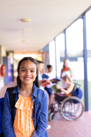 Portrait of smiling biracial schoolgirl standing with bag in school corridor. Education, inclusivity, elementary school and learning concept.の写真素材