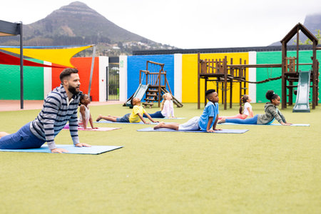 Diverse male teacher and elementary schoolchildren practicing yoga in schoolyard. Education,...