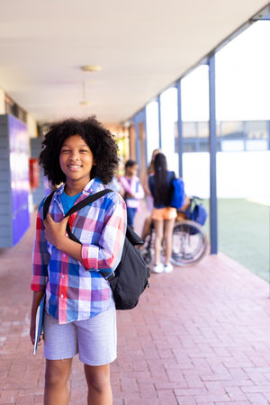 Portrait of smiling biracial schoolgirl standing with bag in school corridor copy space. Education, inclusivity, elementary school and learning concept.の写真素材