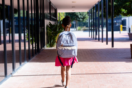 Rear view of biracial schoolgirl with school bag walking in corridor outside school, with copy space. Education, inclusivity, childhood, elementary school and learning concept.の写真素材
