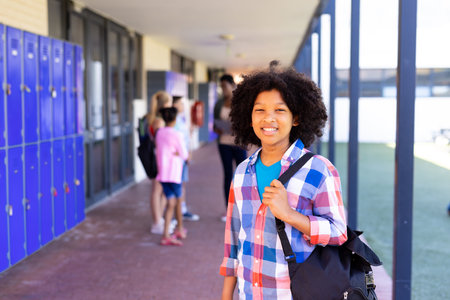 Portrait of smiling biracial schoolgirl standing with bag in school corridor with copy space. Education, inclusivity, elementary school and learning concept.の写真素材