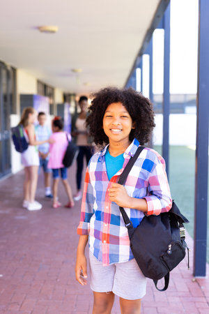 Portrait of smiling biracial schoolgirl standing with bag in school corridor, copy space. Education, inclusivity, elementary school and learning concept.の写真素材