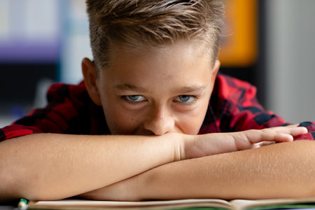 Portrait of tired, bored caucasian schoolboy at desk, resting head on arms in class. Education,...