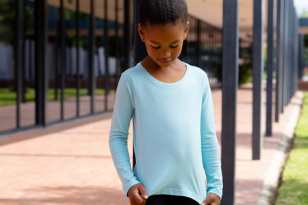Biracial schoolgirl smiling, looking down at her top in sunny schoolyard copy space. Education, inclusivity, childhood, elementary school and learning concept.の写真素材