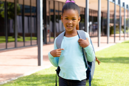 Portrait of happy biracial schoolgirl with school bag smiling in schoolyard. Education, inclusivity, childhood, elementary school and learning concept.の写真素材