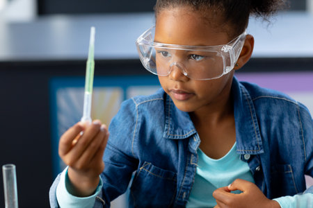 Happy biracial schoolgirl in safety glasses observing pipette in science class with copy space....
