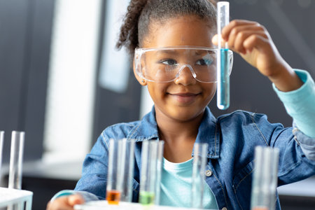 Smiling biracial schoolgirl in safety glasses holding test tube in science class with copy space. Education, inclusivity, childhood, elementary school and learning concept.の写真素材