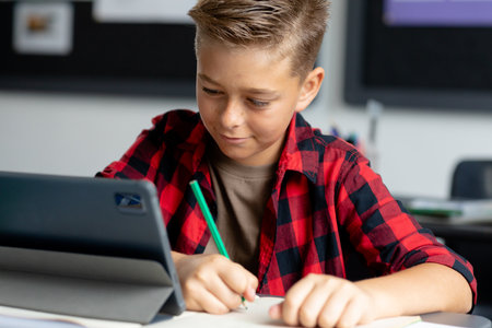 Happy caucasian schoolboy sitting at desk using tablet in class and writing. Education, childhood, elementary school and learning concept.の写真素材