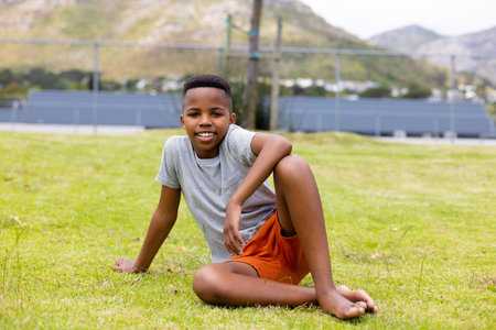 Portrait of happy african american schoolboy sitting on grass at school. School, education and learning concept.の写真素材