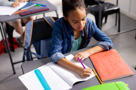 Happy biracial schoolgirl sitting at desk and taking notes in school classroom. School, education and learning concept.の写真素材