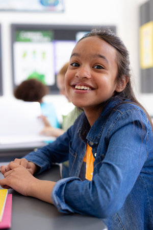 Portrait of happy biracial schoolgirl sitting at desk in school classroom. School, education and...